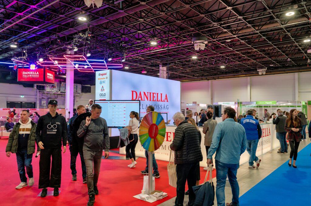Stánek společnosti DANIELLA na veletrhu CONSTRUMA v HUNGEXPO Budapest, kde návštěvníci sledují prezentaci elektroinstalačních technologií a interaktivní program.