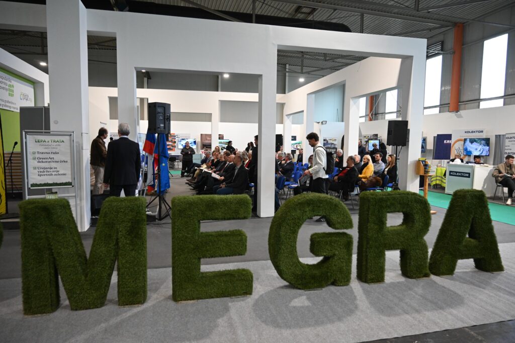 Interiér veletrhu MEGRA s živou prezentací a návštěvníky, v popředí velké travnaté 3D písmeno nápisu MEGRA, stánky vystavovatelů a odborný program.