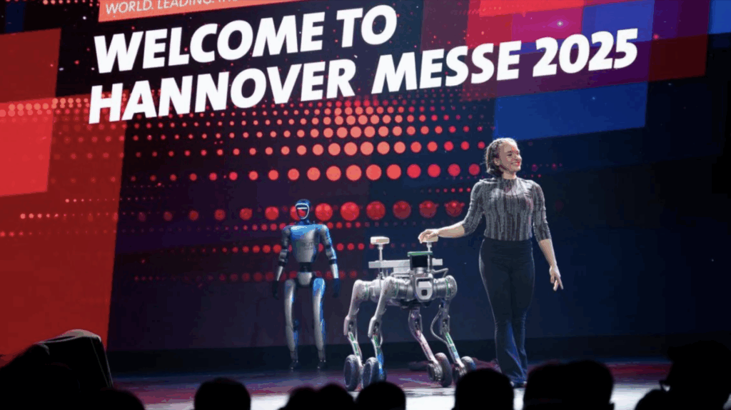Ukázka robotiky na pódiu Hannover Messe, kde robotický pes a humanoidní robot předvádějí technologické dovednosti.