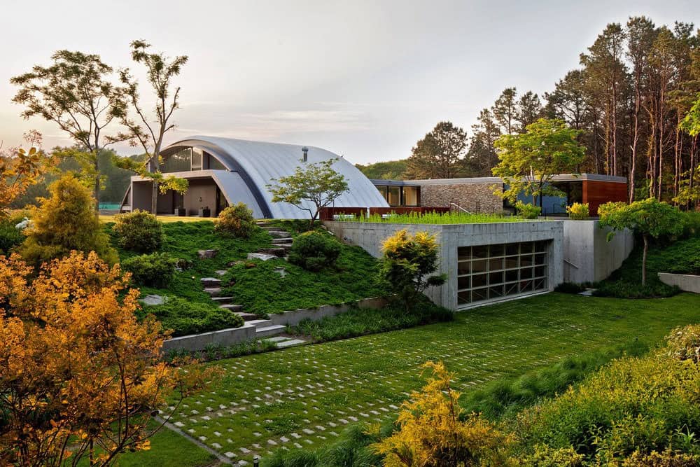 Moderní oblouková ocelová hala integrovaná do svažité zahrady s terénními úpravami, zelení a architektonickými prvky. Ukázka elegantního a funkčního řešení hal v přírodním prostředí.