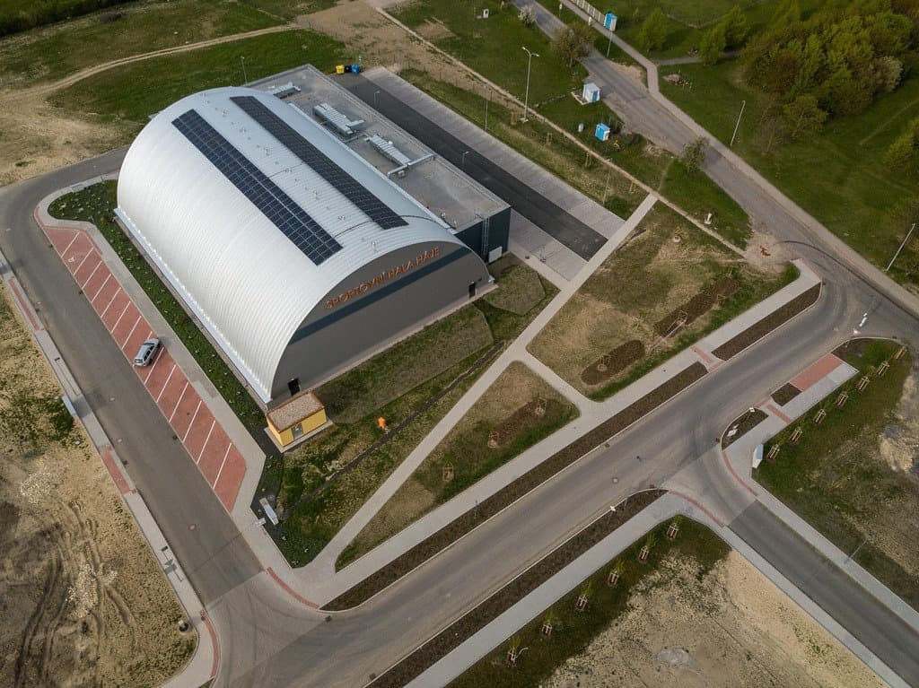 Letecký pohled na obloukovou montovanou sportovní halu HUPRO s okolní infrastrukturou a parkovacími plochami.