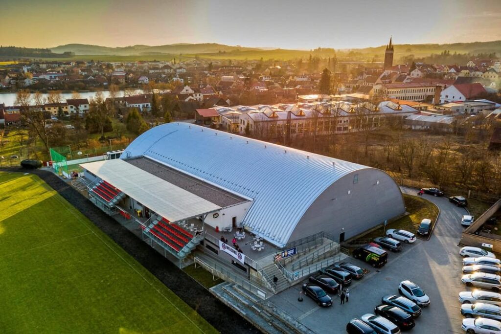 Oblouková sportovní hala HUPRO u hokejového hřiště v Telči s tribunou, parkovištěm a okolní zástavbou.